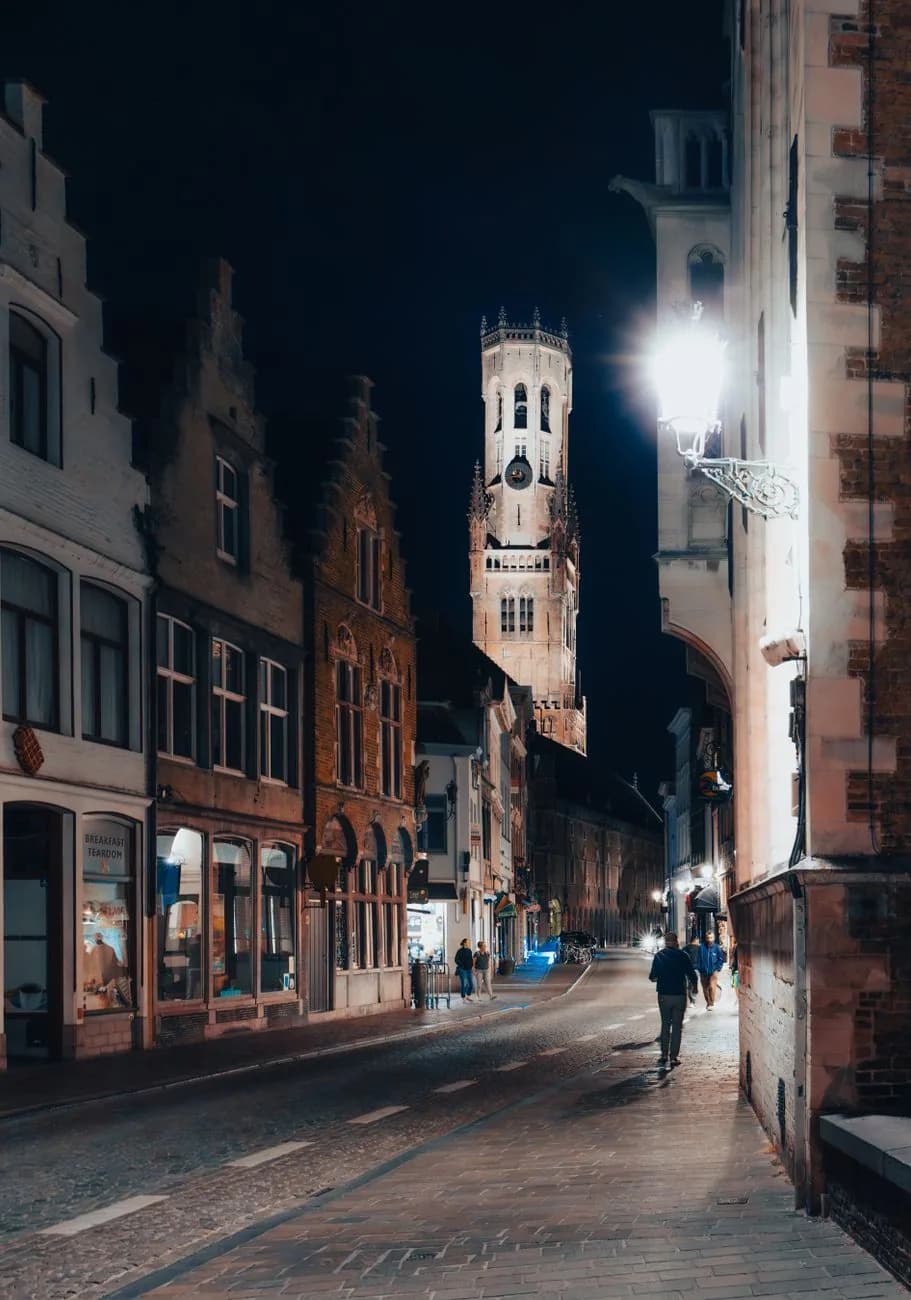 Le beffroi de Bruges illuminé la nuit, au bout d'une rue pavée bordée de maisons à pignons