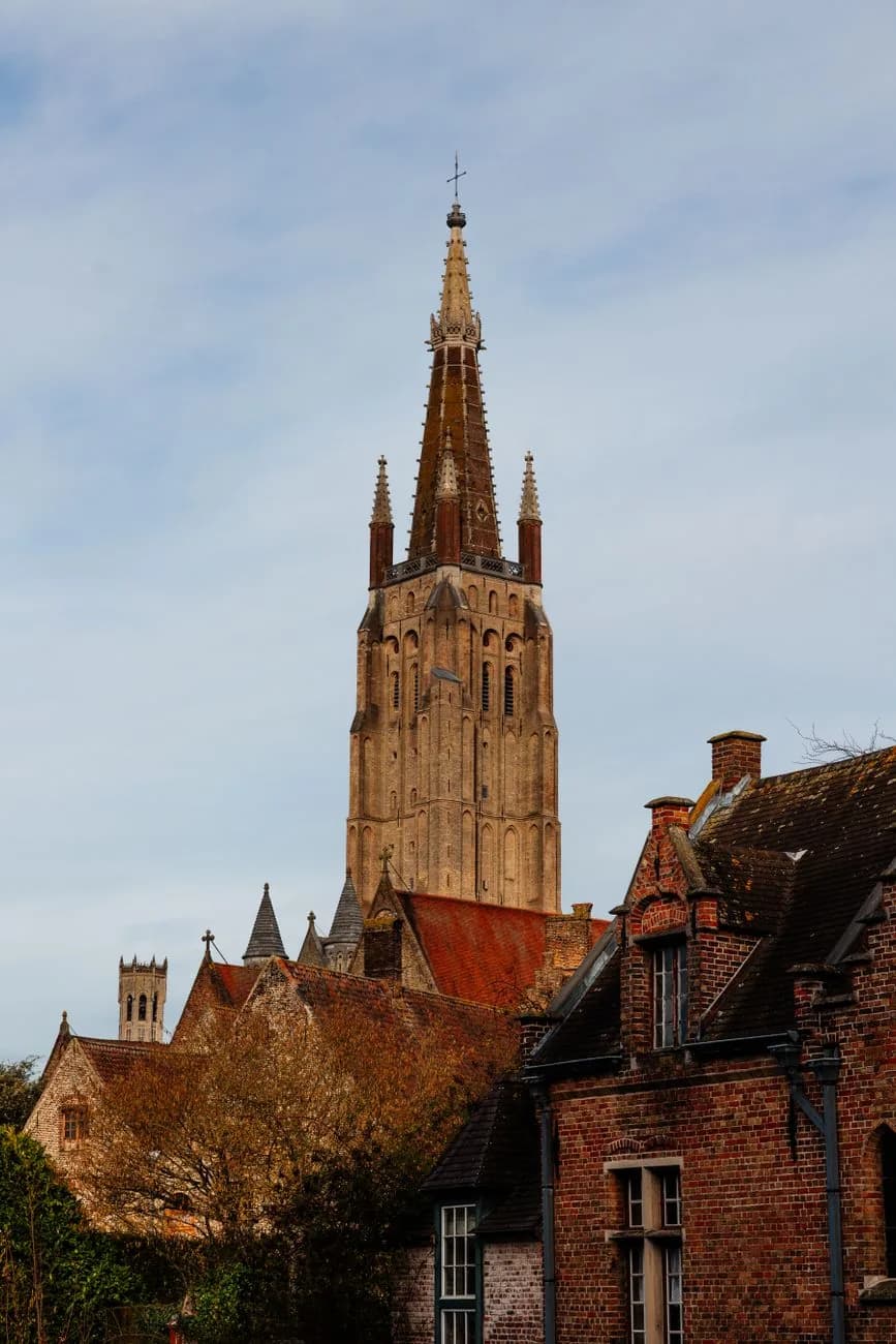 La haute flèche en brique de l'église Notre-Dame de Bruges dominant des toits de tuiles, le beffroi au loin