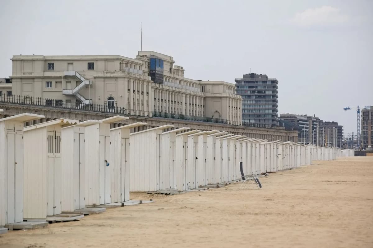 Rangée de cabines de plage blanches sur le sable, devant une digue bordée d'un long bâtiment à colonnades et d'immeubles élevés