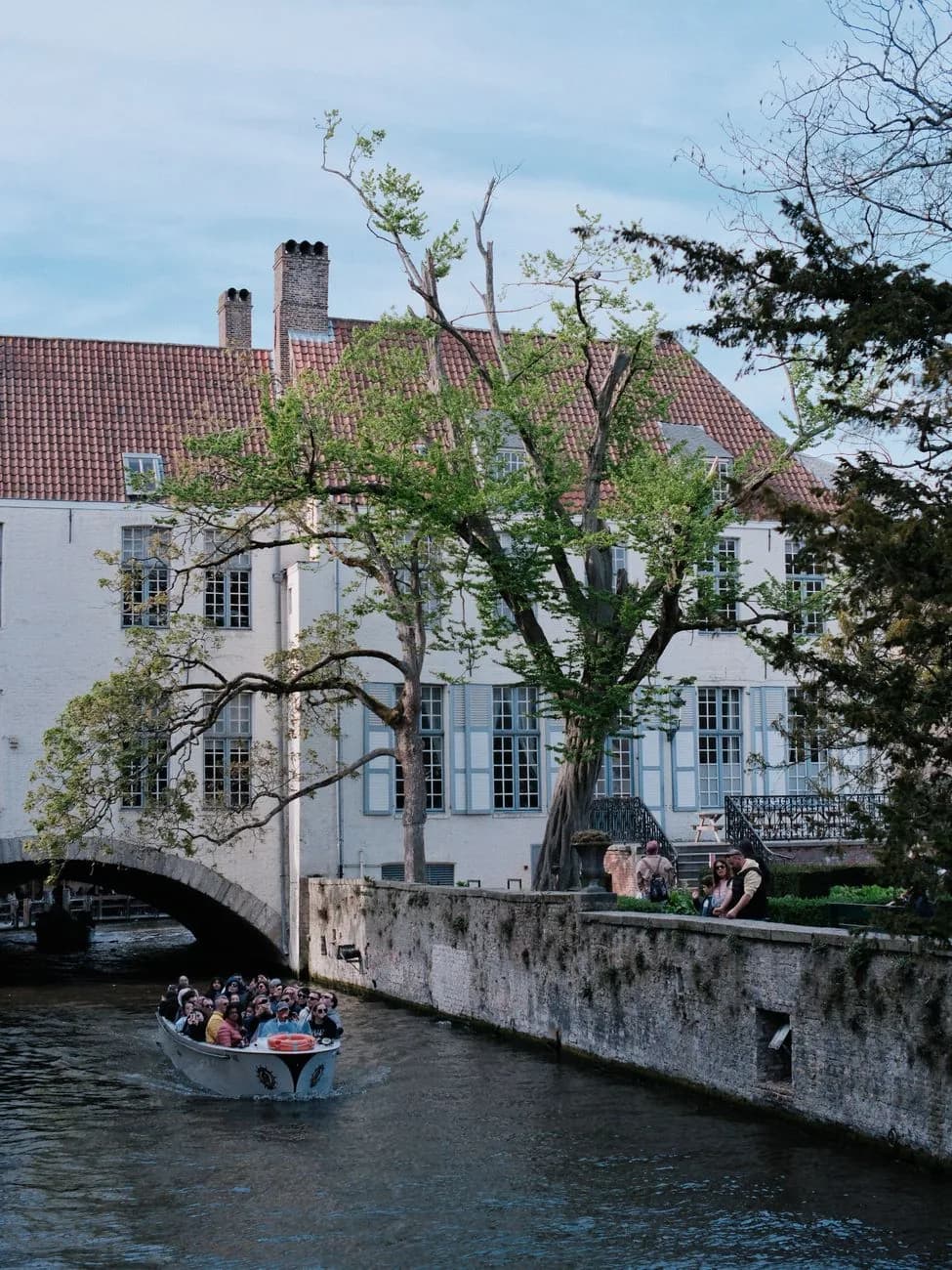 Bateau de promenade passant sous un pont de pierre, devant un bâtiment blanc aux volets bleus au bord de l'eau