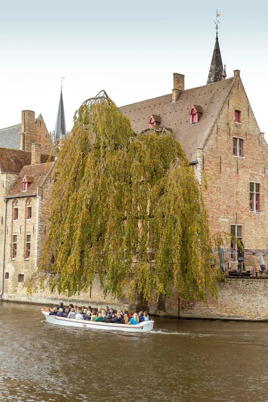 Bateau de promenade rempli de passagers sur un canal, devant une maison ancienne en brique et un saule pleureur