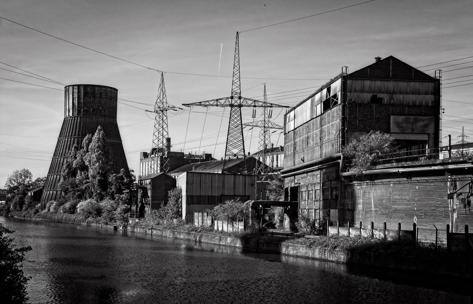 Tour de refroidissement envahie par la végétation et halles industrielles au bord de la Sambre à Charleroi, en noir et blanc