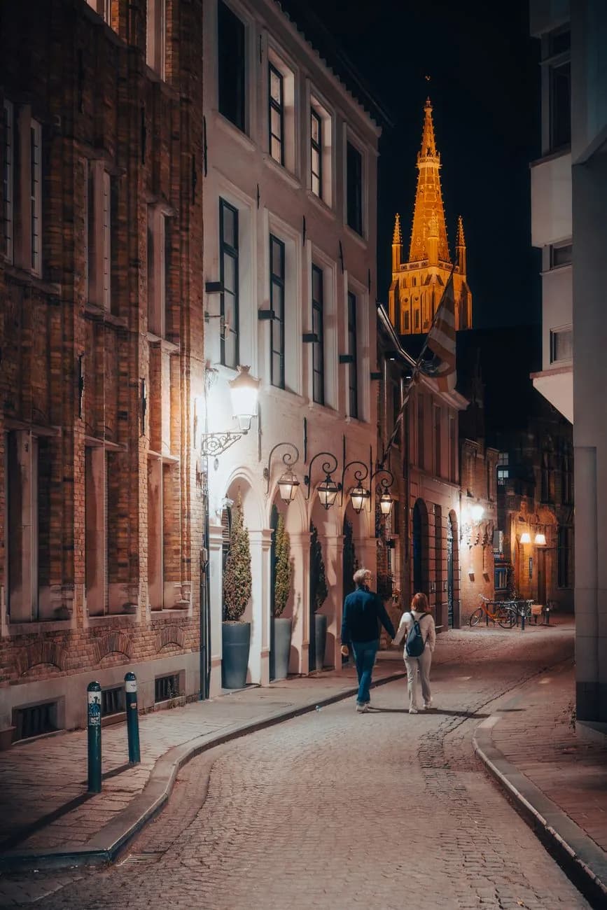 Un couple marche dans une rue pavée la nuit, sous un clocher gothique illuminé