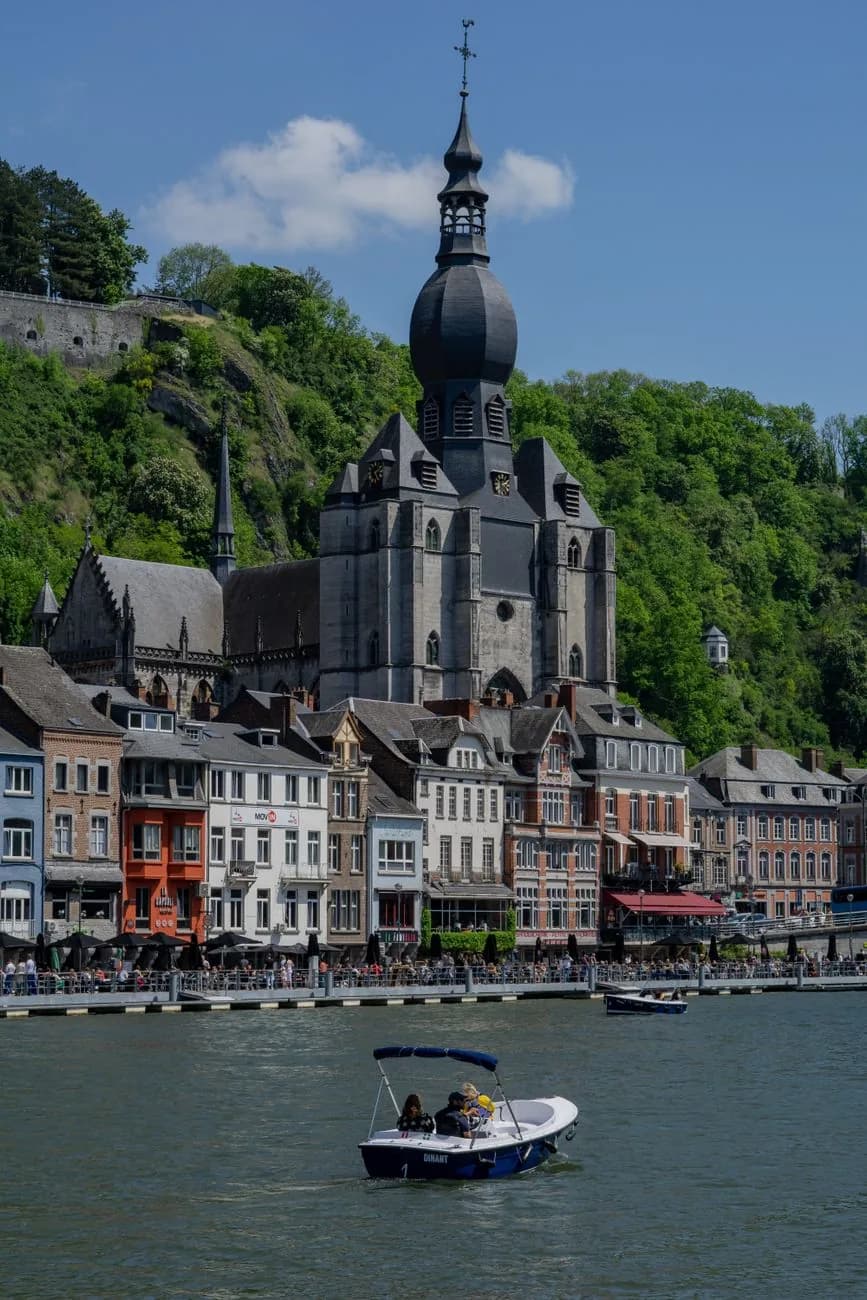 La collégiale Notre-Dame de Dinant et son clocher à bulbe noir, au-dessus des façades du quai, avec un petit bateau sur la Meuse au premier plan