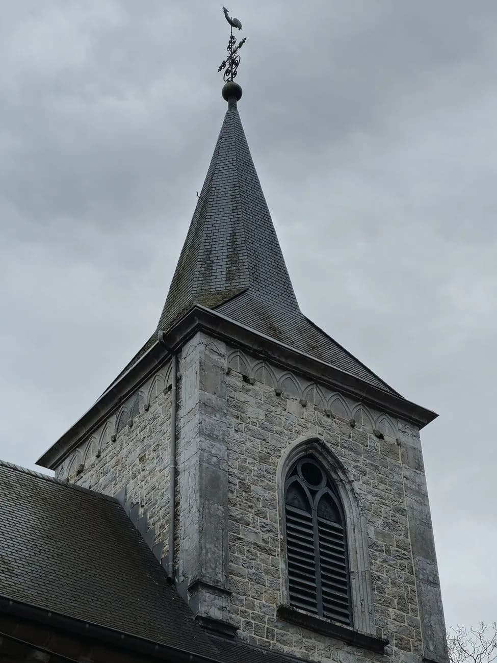 Clocher en pierre d'une église, sa flèche couverte d'ardoises et son coq, sous un ciel gris