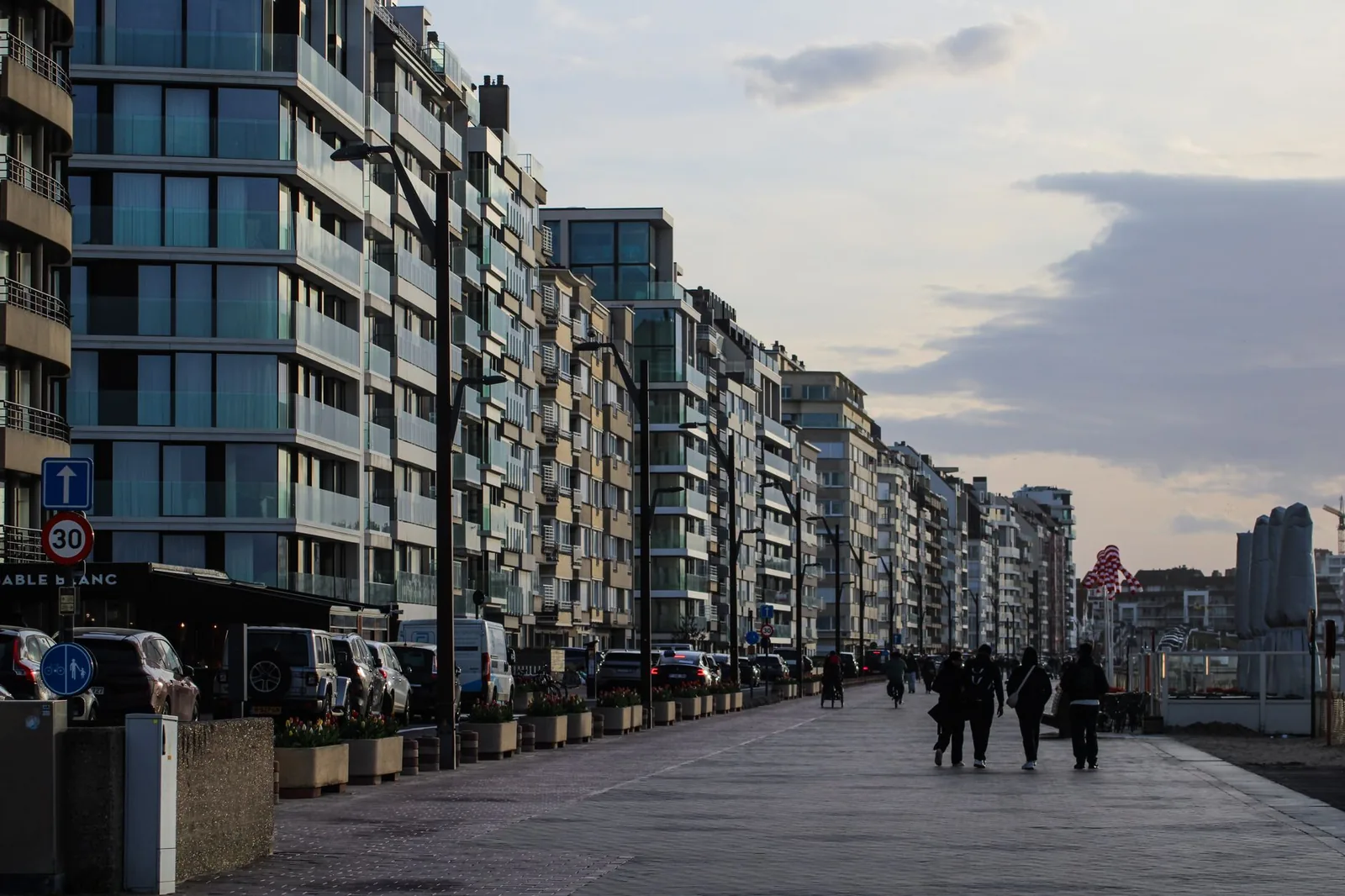 Promeneurs sur la large digue piétonne de Knokke au crépuscule, le long d'une rangée continue d'immeubles à balcons vitrés