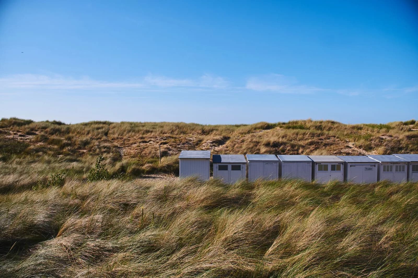 Cabines de plage blanches alignées au pied d'une dune couverte d'oyats, sous un ciel bleu