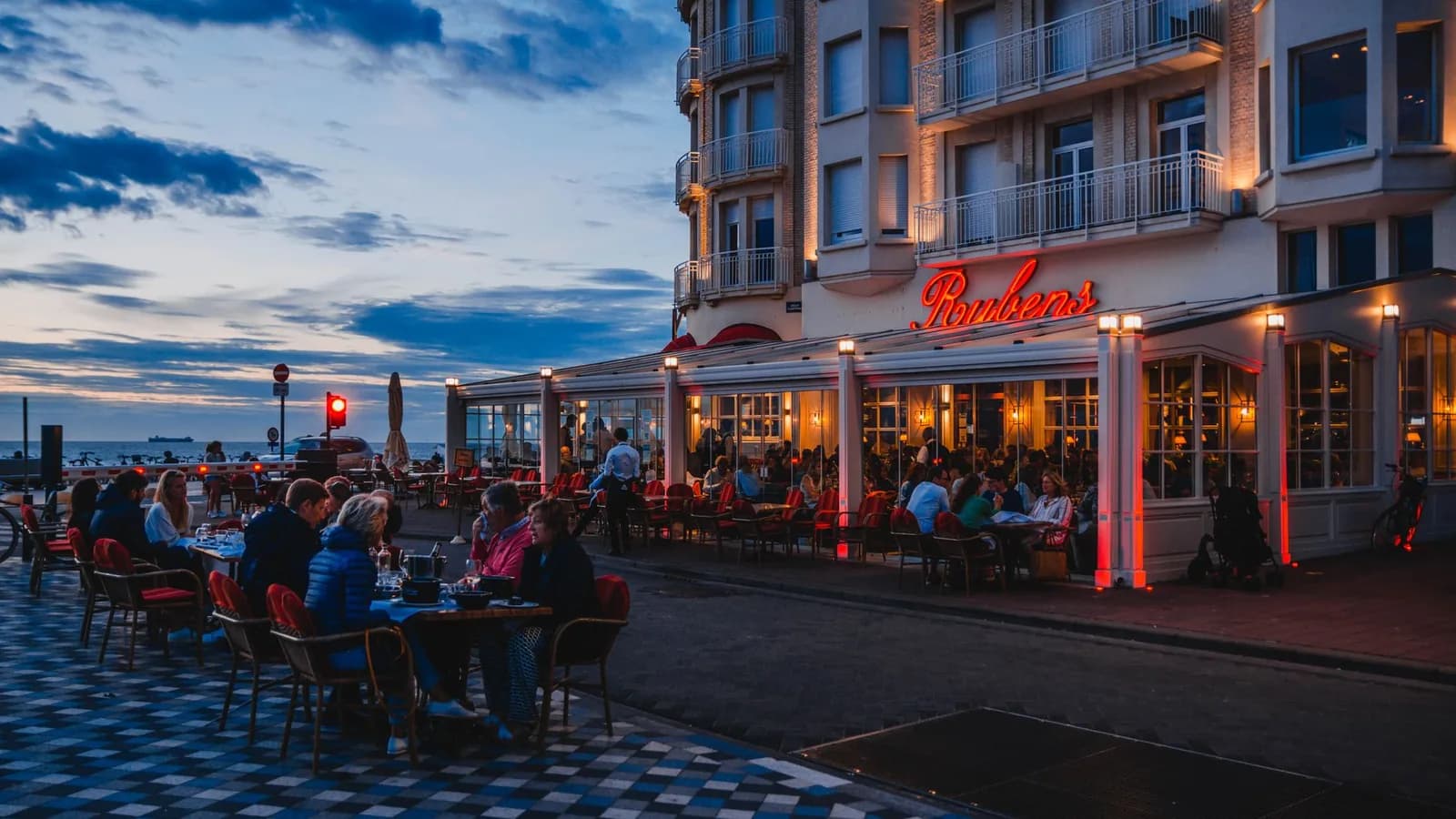 Terrasse pleine d'un restaurant du front de mer de Knokke à la tombée du jour, enseigne lumineuse rouge et mer à l'horizon