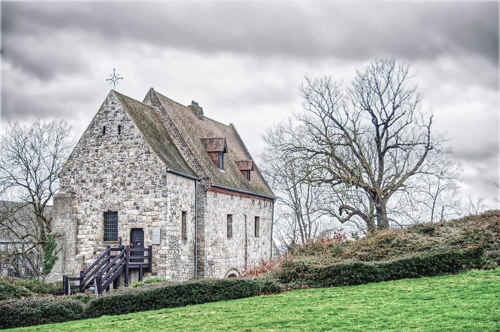 Petite chapelle de pierre au toit pentu, sur la pelouse d'un parc de Mons, sous un ciel gris