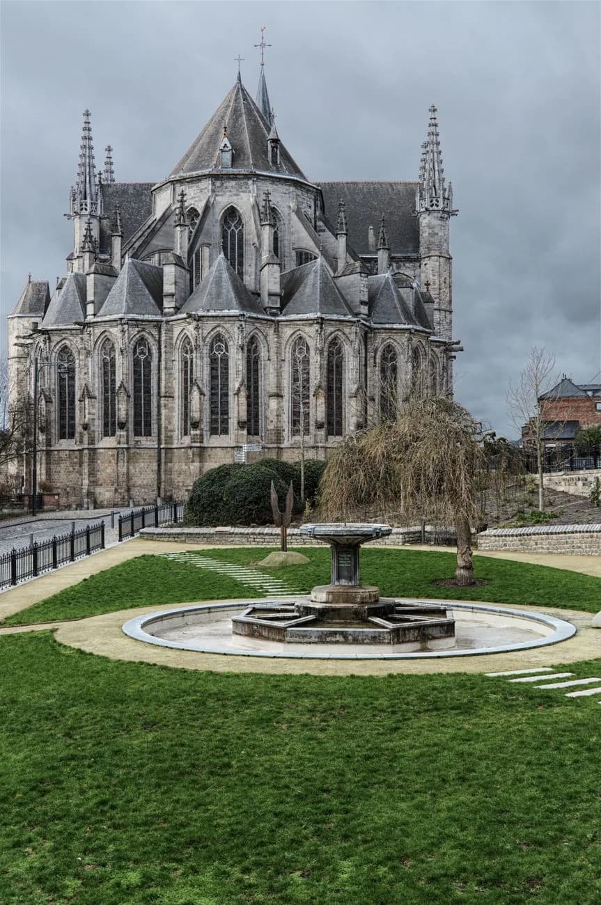 Le chevet gothique d'une grande église de pierre à Mons, derrière une fontaine circulaire posée sur une pelouse