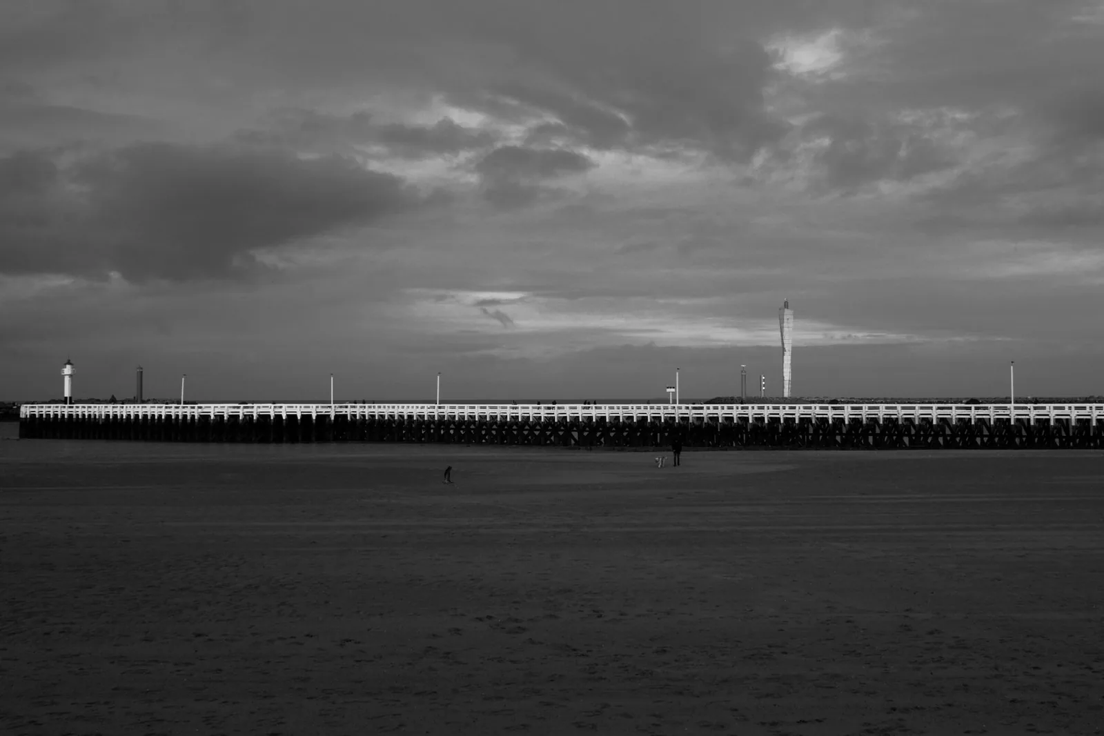 Longue estacade en bois à balustrade blanche prolongée d'un petit phare, sur une plage déserte, en noir et blanc