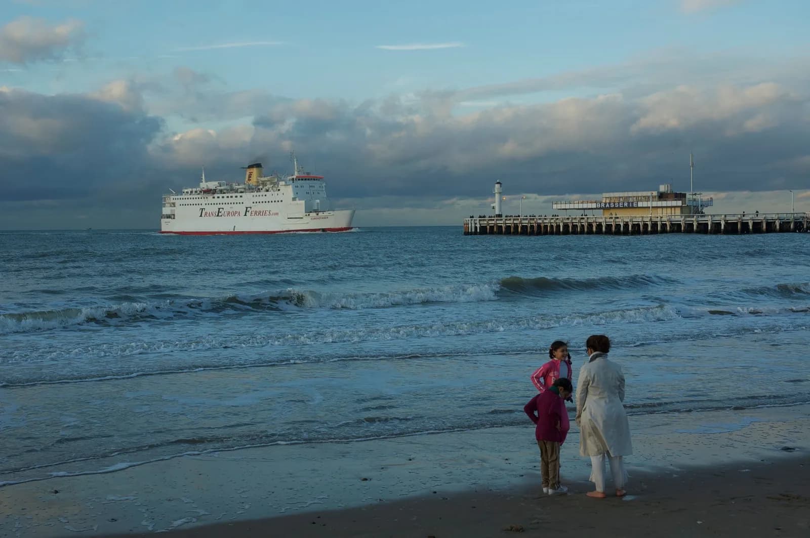 Une femme et deux enfants au bord de l'eau regardent un ferry quitter le port d'Ostende, devant la jetée ouest et son phare