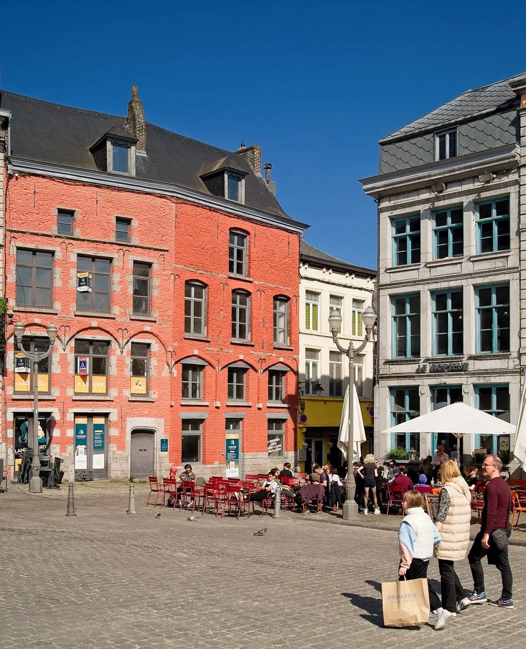 Terrasse de café sur une place pavée de Mons, devant une maison de brique rouge, sous l'enseigne du Ropieur