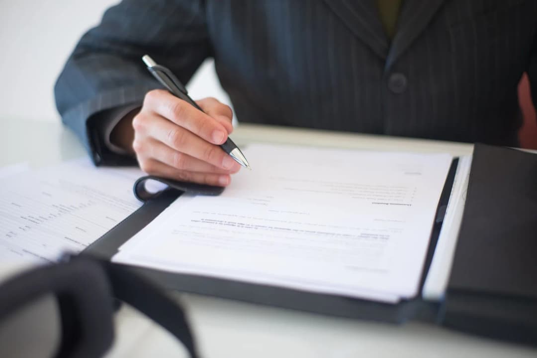 Une personne en costume signe un contrat posé dans un porte-documents, sur un bureau