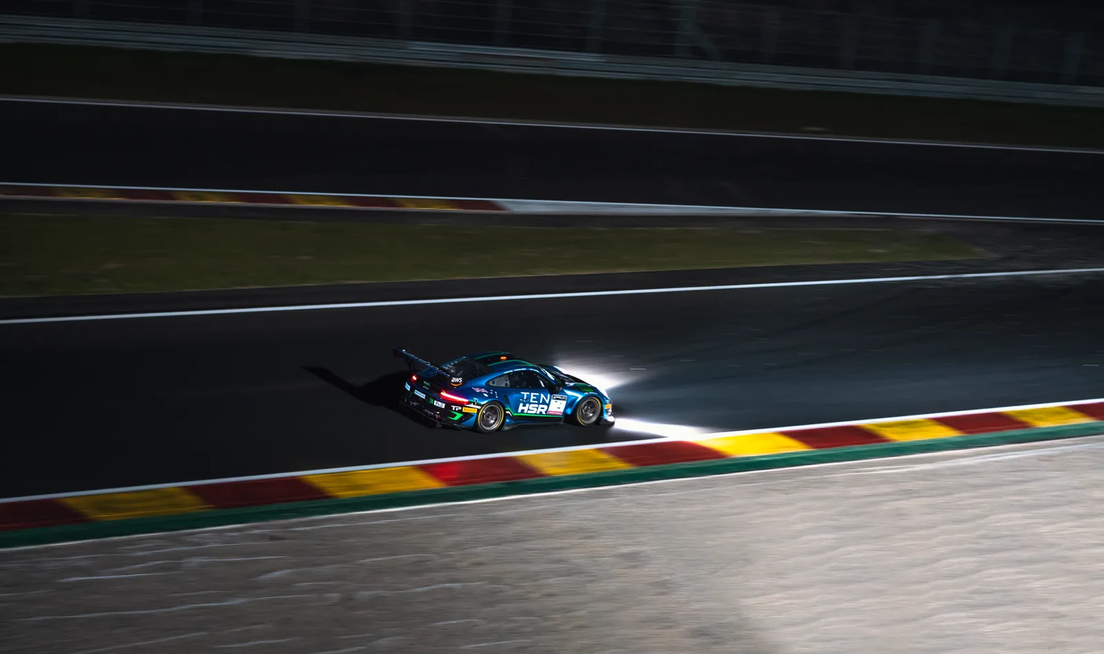 Une voiture de course GT, phares allumés, sur l'asphalte sombre du circuit de Spa-Francorchamps le long des vibreurs rouge et jaune