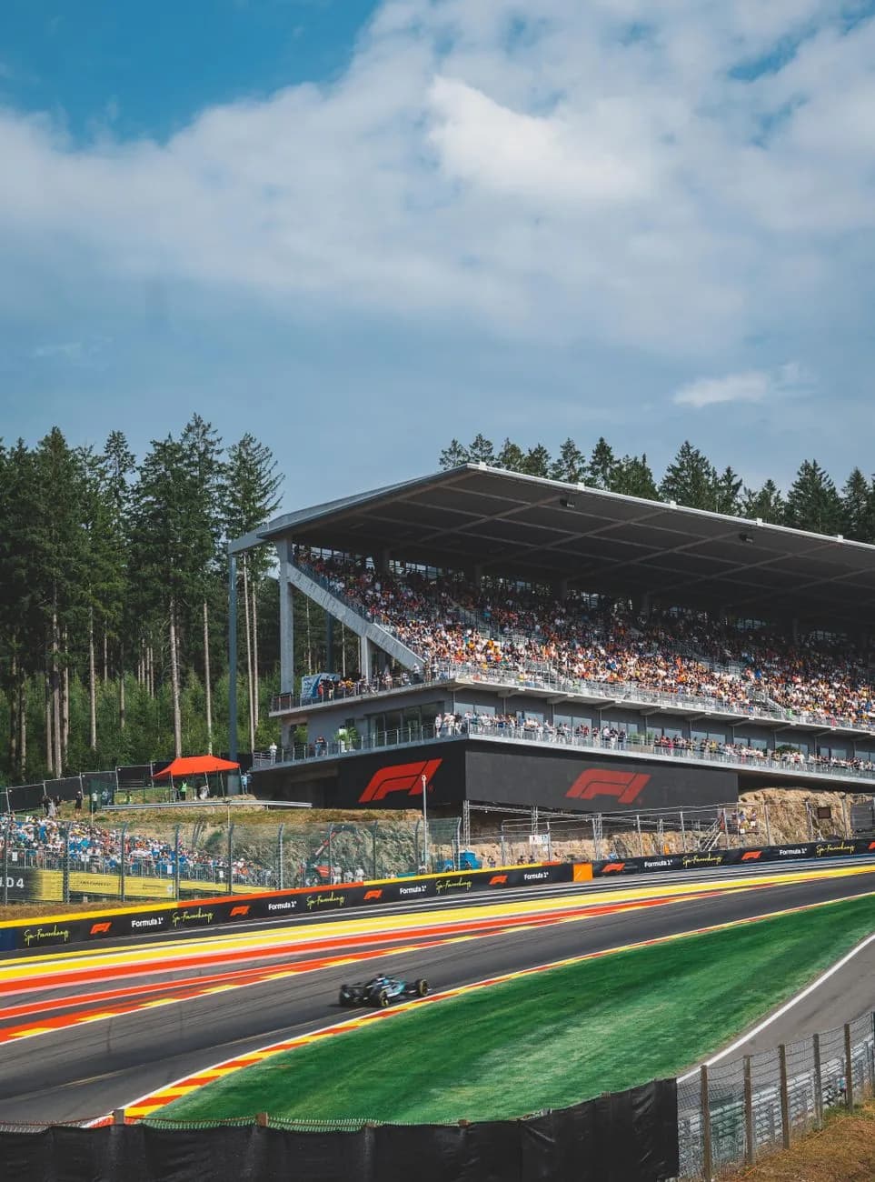 Une Formule 1 passe devant la grande tribune du circuit de Spa-Francorchamps, entre sapinière et vibreurs rouge et jaune