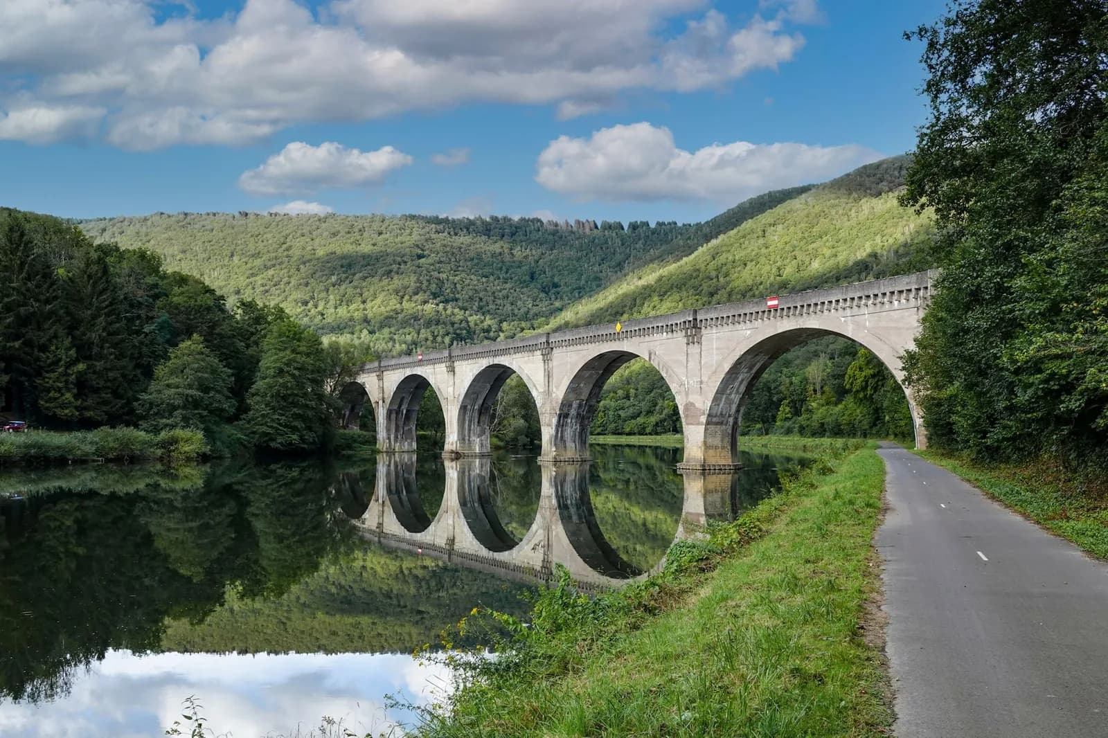 Viaduc en pierre à arches se reflétant dans une rivière calme, bordé d'une piste cyclable au pied de collines boisées